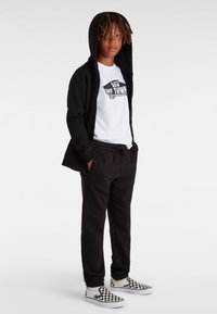 Svart zip-hoodie över en vit grafisk T-shirt, tillsammans med svarta sweatpants och rutiga slip-on skor. Avslappnad outfit med huva.