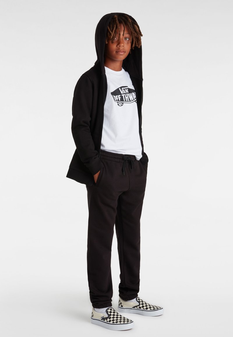 Svart zip-hoodie över en vit grafisk T-shirt, tillsammans med svarta sweatpants och rutiga slip-on skor. Avslappnad outfit med huva.