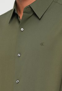 Πουκάμισο σε ελιά πράσινο, κατασκευασμένο από απαλό ύφασμα, με γιακά τύπου spread, ασημί κουμπιά και μικρό ανάγλυφο λογότυπο στο στήθος.