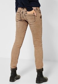 Jean slim marron avec une texture lisse, présentant des poches arrières cousues et des ourlets retroussés sur des bottines noires.