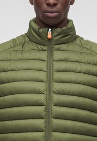 Gros plan d’une personne portant une veste matelassée vert olive avec un col montant et une fermeture éclair avant avec une tirette orange.