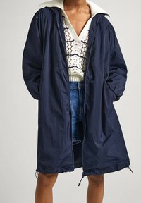 Člověk oblečený v tmavě modré parce s kapucí přes bílý háčkovaný svetr s V-výstřihem a džínové kraťasy, stojící s rukama v kapsách.