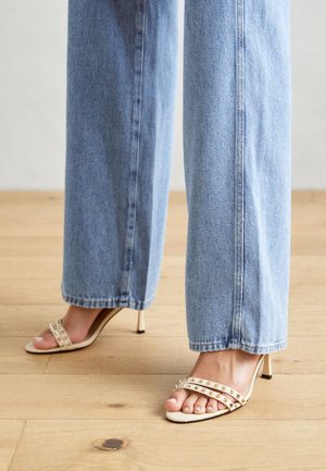 Barna magas sarkú szandál két vízszintes pánttal, amelyek fém tüskék díszítik. Lazán illeszkedő világoskék farmernadrággal párosítva.