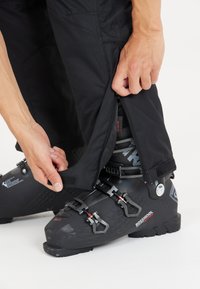 Zwarte skibroek met ritsen aan de onderkant, verstelbare pasvorm en klittenbandsluitingen, gecombineerd met zwarte skibotten met gespen en een gestructureerd oppervlak.