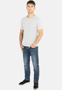 Weißes Rundhals-T-Shirt mit kurzen Ärmeln, kombiniert mit blauen Jeans. Modell trägt schwarze sportliche Sneakers und steht vor einem schlichten Hintergrund.