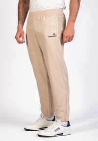 Pantalones de chándal beige con cinturilla elástica, bolsillos laterales y detalle de cremallera en los puños, con un logotipo oscuro. Llevados con zapatillas blancas.