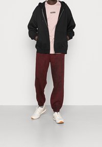 Sort hættejakke med lynlås, lys pink t-shirt med logo, bordeaux sweatpants med elastiske manchetter og hvide træningssko med orange detaljer.