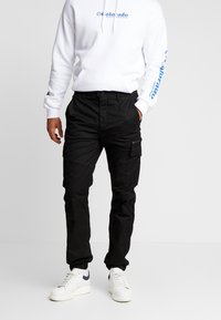 Zwarte cargobroek met meerdere zakken en een relaxte pasvorm, gecombineerd met een witte hoodie met blauwe tekst en logo-accenten.