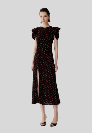 Donna che indossa un vestito midi nero con stampa floreale rossa, maniche a sbuffo, spacco laterale e tacchi neri aperti, in piedi davanti a uno sfondo semplice.