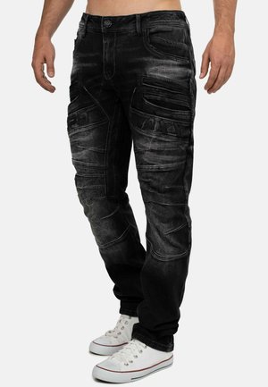 Black faded jeans with stitched panel details and embossed text, worn with white low-top sneakers on a person standing.