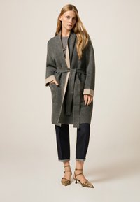Langer grauer Cardigan mit beigen Akzenten, tailliert. Wird über einem grauen Top und dunklen Jeans getragen, ergänzt durch High Heels mit Schlangenprint.