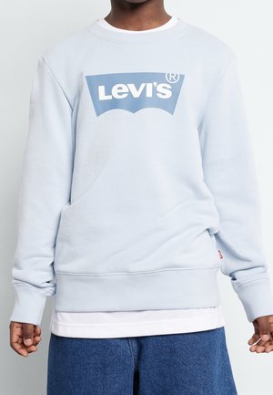 Personne à la peau foncée portant un sweat-shirt Levi's bleu clair par-dessus une chemise blanche et un jean bleu, debout devant un fond uni.