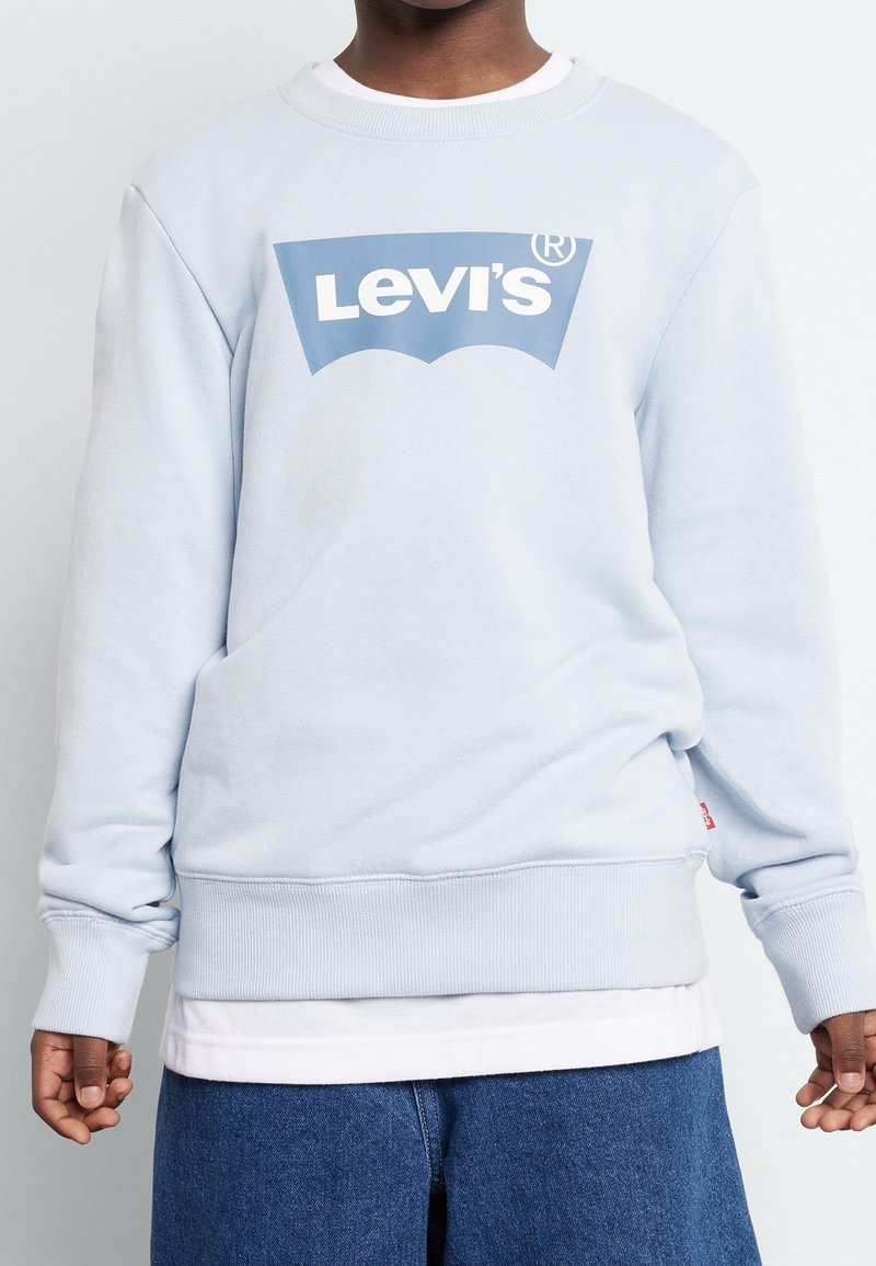 Personne à la peau foncée portant un sweat-shirt Levi's bleu clair par-dessus une chemise blanche et un jean bleu, debout devant un fond uni.