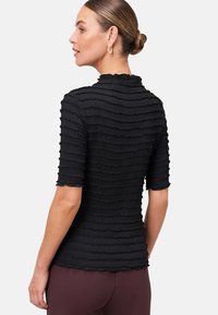 Donna con capelli raccolti in uno chignon, indossa una maglietta nera a maniche corte con texture e pantaloni bordeaux, rivolta verso sinistra su uno sfondo bianco semplice.