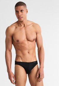 Schwarze Bikiniunterwäsche aus glattem Stoff, mit einem niedrigen Schnitt, minimaler Naht und einem kleinen Logosticker am Bund.
