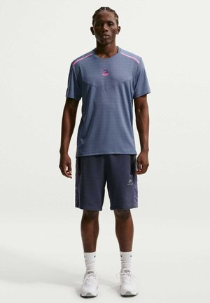 Homme debout portant un maillot de sport Nike bleu, un short foncé, des chaussettes blanches Nike et des chaussures de sport blanches sur un fond uni.