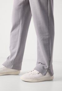 Pantalones grises claros hechos de tela suave, con un zipper lateral en el tobillo, combinados con zapatillas blancas. Diseño simple de pierna recta.