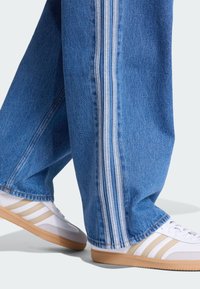 Pantaloni di denim azzurri con tre strisce verticali bianche lungo i lati, abbinati a scarpe da ginnastica bianche con dettagli beige e suole in gomma.
