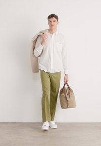 Camicia bianca leggero con bottoni, pantaloni verde oliva e sneakers bianche. Porta una borsa da viaggio color beige con dettagli marroni. Maglione beige sulle spalle.