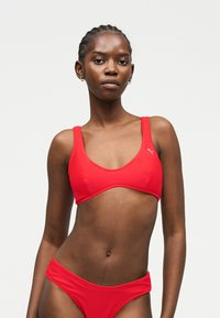 Mujer con el pelo trenzado que lleva un conjunto de bikini rojo de Puma, de pie frente a un fondo claro y liso, mirando al frente con expresión neutra.