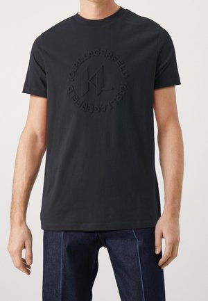 Sort bomulds T-shirt med korte ærmer, der har et prægget logo og cirkulært tekstdesign på forsiden.