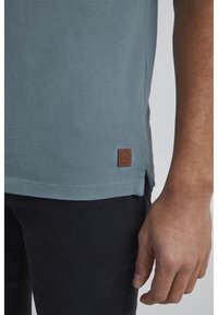 Lyseblå polo-skjorte med et struktureret stof, der har et brunt mærke ved kanten. Skjorten er parret med mørke bukser.
