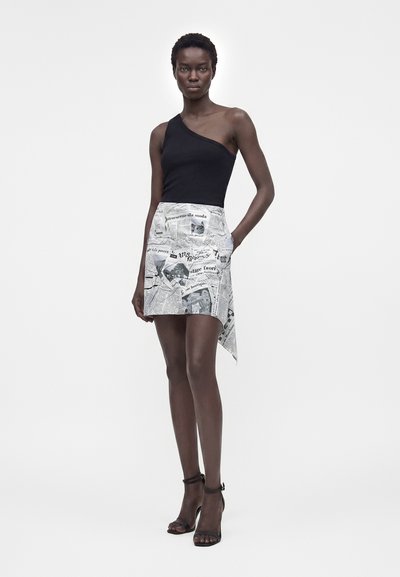 Γυναίκα με σκούρο δέρμα που φοράει μαύρο τοπ με έναν ώμο, ασύμμετρη μίνι φούστα με τύπωμα εφημερίδας και μαύρα πέδιλα με λουράκι στον αστράγαλο, στέκεται με το ένα χέρι στην τσέπη.