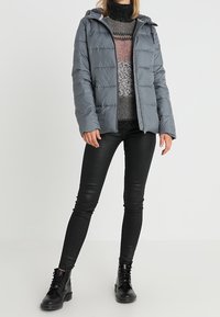 Grå puffjacka med huva, quiltad design, som bärs över en flerfärgad mönstrad tröja. I kombination med svarta tajta jeans och kängor.