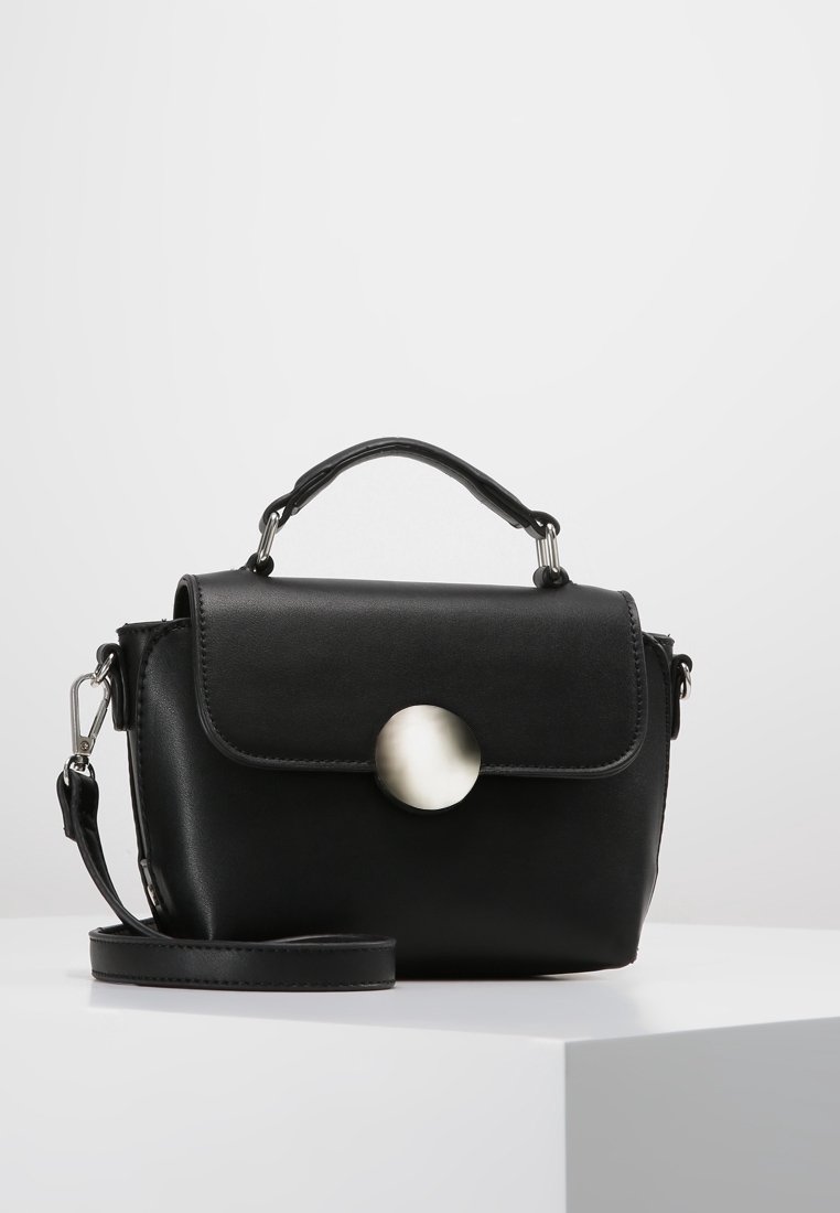 Borsa a mano in pelle nera con manico superiore, chiusura a patta caratterizzata da un grande pulsante rotondo in argento, e tracolla rimovibile appoggiata su una superficie bianca.