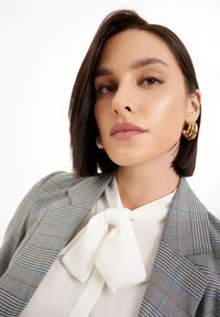 Blazer de cuadros grises con acentos en azul y naranja, blusa blanca con lazo y grandes pendientes de aro dorados. Cabello liso y pulido.