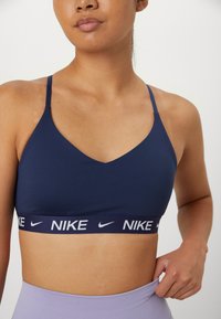 Marine sports-bh med tynne skulderstropper og v-hals design. Har en hvit Nike-logo på det elastiske båndet. Glatt tekstur, tettsittende fasong.