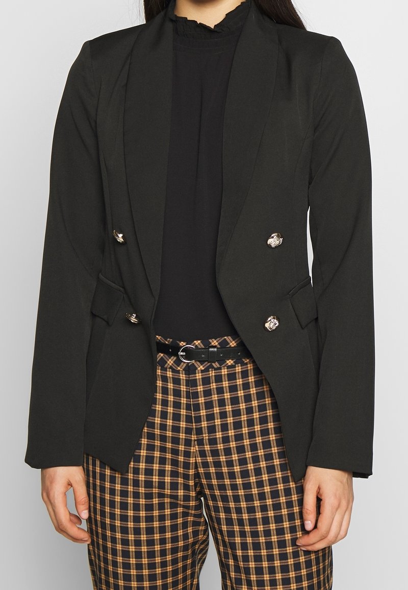 Femme portant un blazer noir croisé à boutons dorés, un chemisier noir à volants et un pantalon marron et noir à carreaux avec une ceinture noire.