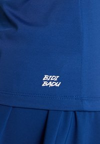 Modré sportovní oblečení s texturovanou tkaninou; má bílé logo "BIDI BADU" poblíž lemu a skládáný design na spodní části.