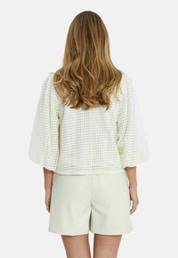 Krämfärgad, strukturerad jacka med 3/4-lång ärm och volangärmar, prydd med ett geometriskt mönster och rak passform, kombinerad med ljusbeige shorts.