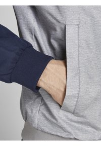 Hand in der Tasche einer hellgrauen Jacke mit dunkelblauem, geripptem Bündchen, sichtbarem Stofftextur und Nahtdetails.