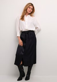 Witte blouse met pofmouwen, gecombineerd met een zwarte satijnen maxi rok met een zijsplit; gedragen met zwarte cowboylaarzen.