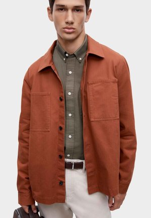 Junger Mann trägt eine orangefarbene Jacke mit Knöpfen über einem olivgrünen Hemd und beigen Hosen, hält eine kleine braune Tasche in einer Hand.