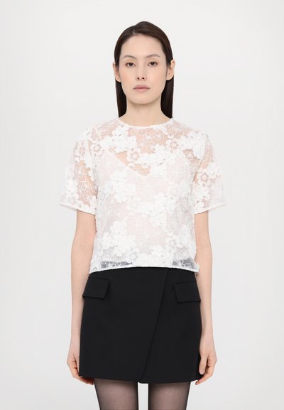Haut en dentelle blanche avec broderie florale, manches courtes et col rond, associé à une jupe noire avec deux poches frontales.