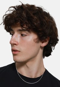Una collana d'argento giace su una maglietta nera, mettendo in risalto un modello maschile con capelli ricci castani e pelle liscia, che volge lo sguardo verso destra.