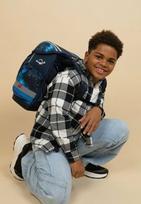 Der Rucksack hat ein navyblaues Äußeres mit einem Galaxiedruck, verstellbaren Riemen und einem reflektierenden Streifen am Boden. Das Kind trägt ein karierte Hemd.