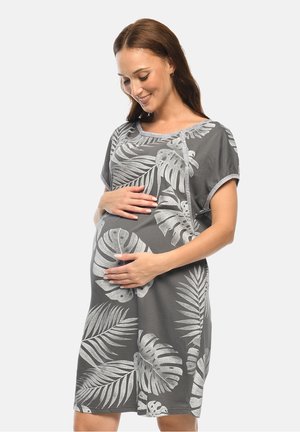 Schwangere Frau, die sanft ihren Bauch hält und ein graues Kleid mit großen weißen tropischen Blattmustern trägt.