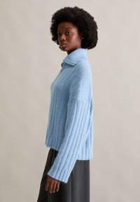 Hellblauer gestrickter Pullover mit kragenförmigem Ausschnitt, geripptem Muster, weiten Ärmeln und lockerer Passform, kombiniert mit einem dunkelgrauen Unterteil.