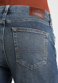 Modré džíny z denimu mají koženou nášivku na zadním pase, jednu zadní kapsu s kontrastním prošíváním a rovný střih.