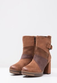 Bottes chelsea en daim marron avec un talon carré, présentant une fermeture éclair et une large sangle décorative. Le design en blocs de couleurs comprend des sections marron foncé et beige.
