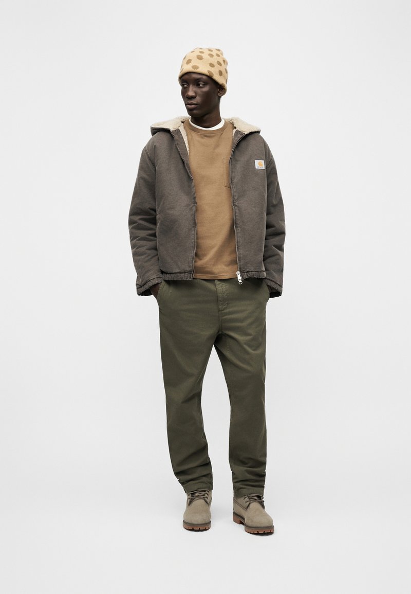 Mann trägt eine braune Mütze mit Punkten, eine braune Jacke mit Schaffellfutter, ein beiges Sweatshirt, olivgrüne Hose und beige Stiefel, steht mit den Händen in den Taschen.