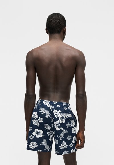 Tume nahaga mees, kes kannab tumesiniseid lühikesi pükse valgete lille- ja leopardimustriga, seisab selg kaamerale heledal ühtlasel taustal.