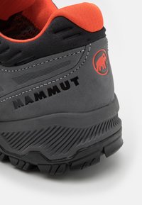 Grå vandresko med orange detaljer, med en tekstureret gummisål, forstærket tå og Mammut-logoet i rødt på hælen.