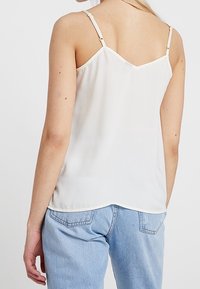 Bílý bezrámový top s tenkými ramínky a výstřihem ve tvaru V na zádech. Vyroben z hladkého materiálu s jemným leskem a volným střihem.