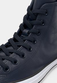 Tenis de înălțime mare, de culoare bleu marin, fabricat din piele texturată, cu șireturi plate, ochiuri metalice și o talpă din cauciuc alb cu un accent negru.