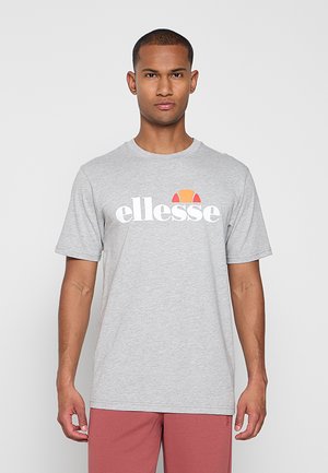 Mann trägt ein hellgraues ellesse-Logo-T-Shirt und rote Shorts, steht vor einem schlichten hellen Hintergrund.